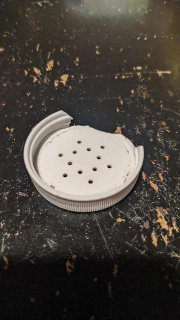 A broken white plastic pepper shaker lid, shattered from falling.