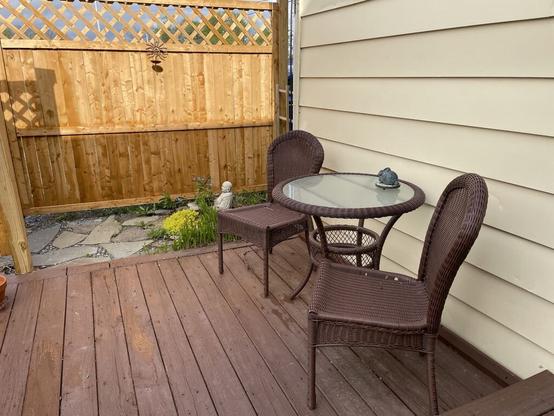 Two wicker chairs are at either side of a matching round-topped table. These are against a yellow exterior wall, on a reddish wooden deck, and backed by a raw wood fence.