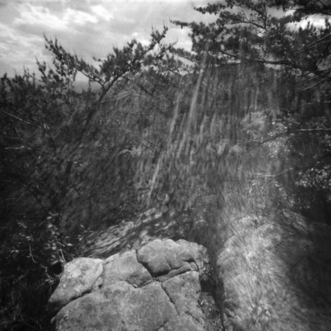 Black and white pinhole photo of a rock ledge and scruffy trees on a mountainside. Much of the scene is covered in light streaks caused by the sun hitting the tiny aperture hole of the camera.