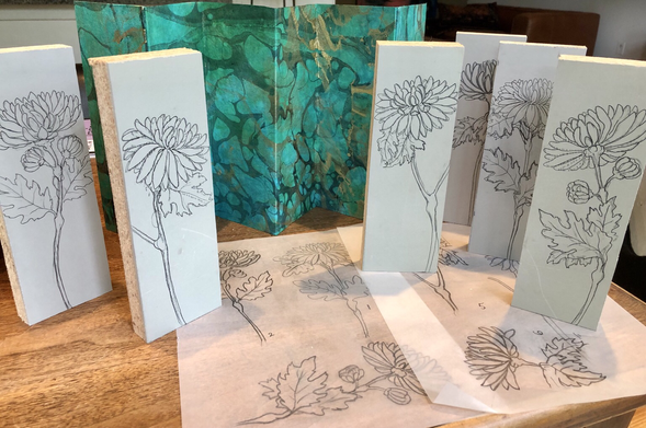 A table top with pencil sketches of 6 different single stem chrysanthemums on 3 by 9 inch linoleum blocks for print making. In the background is an 18 by 10 inch folding screen with green marbled paper.