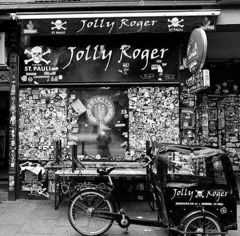 Black and white photo, outside front of bar in Hamburg,  covered with stickers with custom bicycle delivery outside.