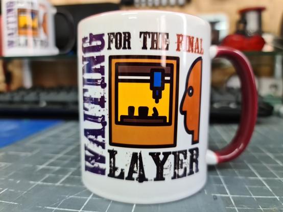 A white printed mug with a maroon handle. The front of the mug has an inconised image of someone looking at a 3d printer, with the words "waiting for the final layer" surrounding it.