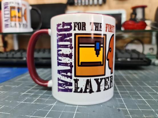 A white printed mug with a maroon handle. The rear of the mug has an inconised image of someone looking at a 3d printer, with the words "waiting for the first layer" surrounding it.