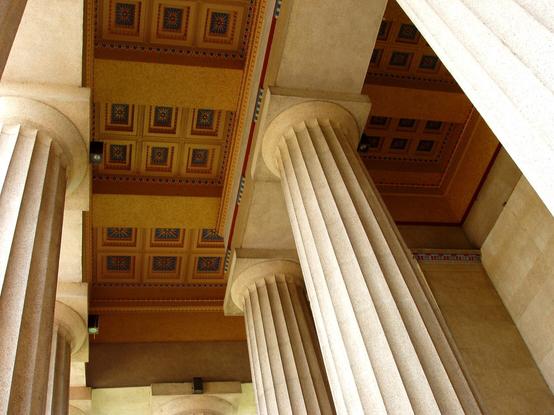 A photo of the east portico of the Nashville Parthenon. We see doric columns ascending to the roof space, where corbelled stones form a chequerboard pattern above the viewer.