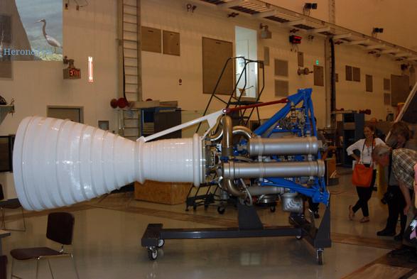A photograph of a rocket engine mounted on a blue frame which rests on a black cart. The top of the engine is on the right, where several reporters and photographers are looking on. Two long tubes reach down the facing side of the engine. The nozzle is made of a white plastic material and is on the left.