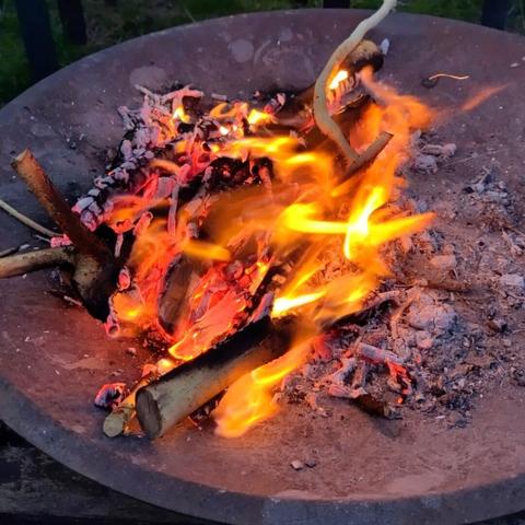An outdoor bonfire of wood burns fiercely in a metal bowl with vibrant orange yellow flames