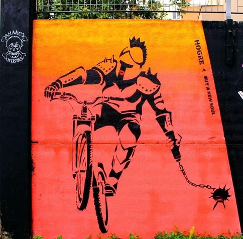 Streetartwall. On an orange-painted street wall, a mural of a black knight on a bike is sprayed in pictogram style. He rides on the bike and swings a steel ball on a chain (a medieval weapon) behind him.