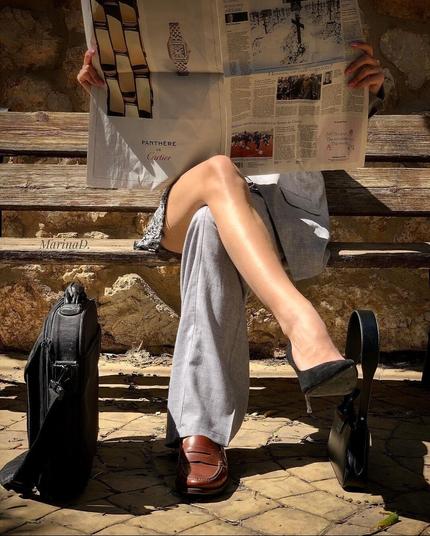 Photography. A color photo of a woman, sitting on a park bench and reading a newspaper. The right leg is crossed over the left leg. On the right she is dressed in short gray skirt, bare legs and black pumps. The left leg, on the other hand, has gray men's suit pants on and a brown men's shoe. On the left is a grey laptop bag on the floor and on the right is a black women's handbag.
"Behind every successful woman is herself"