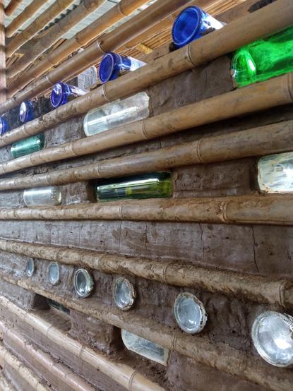 A bamboo and mud wall with green and clear glass bottles encrusted within it.