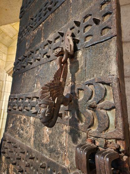 Ancient fortified door with a knocker in the shape of a mystical figure with body of a bird(?) and head of a person wearing a headdress of feathers.