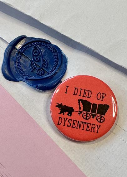 A pin that says “I died of dysentery” and shows the wagon from Oregon Trail game on top of an envelope with a wax seal