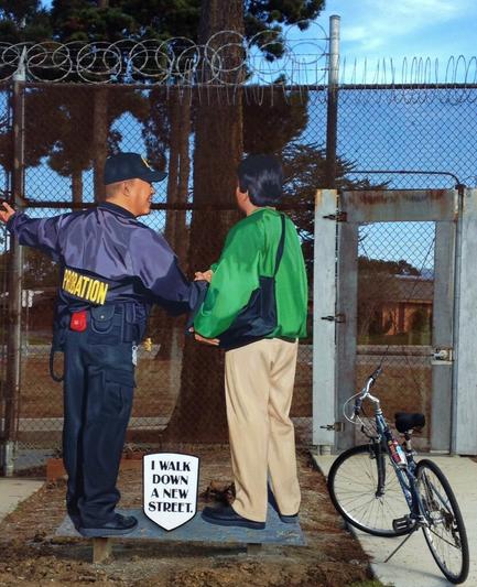 A mural by artist John Cerney is at the entrance/exit gate of the Monterey County Probation Department Youth Center. The wire fence alongside the gate is topped by razor wire. The mural shows a probation officer standing alongside a teen boy. The officer's hand is outstretched, indicating to the boy the world that awaits him. A sign at their feet says "I walk down a new street." A bicycle alongside the mural expresses "I bike down a new street" too!