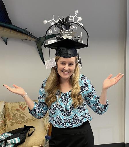 Nicely-dressed woman with blond hair wearing a giant graduation cap highlighting her projects and interests smiles at the camera following her successful thesis defense