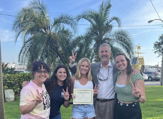 Four graduate students and the Dean pose looking at the camera on a gorgeous Florida day, palm trees in the background. They are making V’s with their fingers in honor of the Vembu award