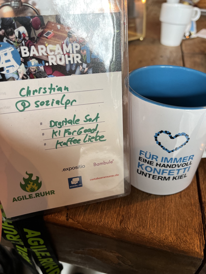 Links mein Namensschild mit dem im Text genannten Hashtags, rechts die Tasse des Barcamps