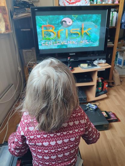 A blond haired boy sitting on a toolbox in front of a TV which shows the opening screen of "Brisk: een levendig spel".
On the floor in front of him is the MSX2 homecomputer that's running the game, with a Philips Music Module and a Panasonic FM-PAC sticking out the side, providing additional music chips for games that support them.
On the shelf on the TV furniture another of the same machines can be seen, as I was experiencing some hardware issues.
A joystick (The Arcade Turbo) can also be seen on the furniture unit.