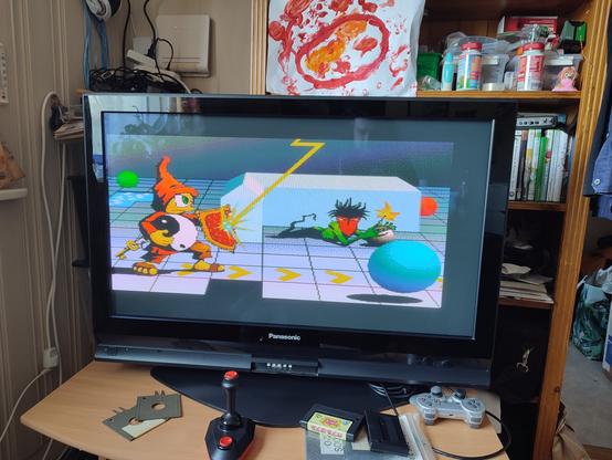 The Loading screen of Brisk; a figure wielding a shield (which is blocking a lightning bolt) and holding a Ying Yang symbol in his other hand, following an arrow path and a ball, while another figure is holding a bomb.
On the TV desk the two transport floppies, a joystick, and the Bubble Bobble cartridge can be seen, as well as MSX DOS 2.20 and 1MB RAM cartridges in a zip lock bag