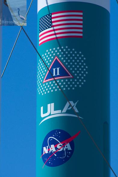A zoomed-in photograph of a teal cylinder with the US flag, A Greek letter Delta, which is a triangle with its point up, and the Roman numerals II inside, with many stars surrounding it. Below that is the ULA logo and then the round NASA blue meatball logo.