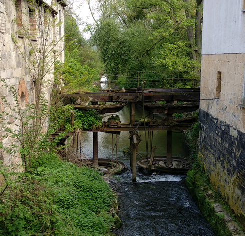 Große Metallräder vom ehemaligen Schaufelrad-Antrieb einer Wassermühle hängen über dem schmalen Wasserlauf eines kleinen künstlichen Kanals.
