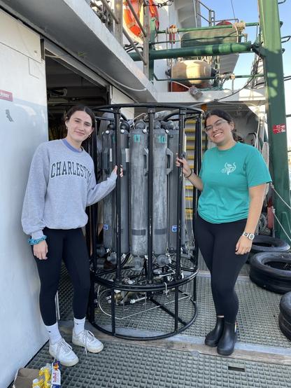 Two women posing by a CTD Rosette on the deck of a research ship