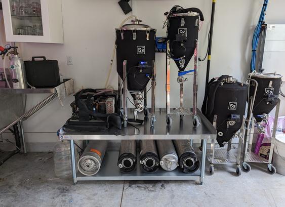 A stainless steel table holding a chiller, 2 fermenting vessels, and 5 kegs