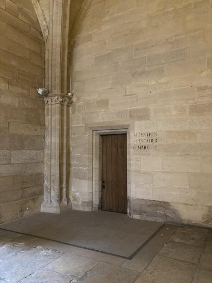 A plain wooden door in a corner of the notorious former prison the Conciergerie is dwarfed by monotone stone walls with the words "SALLE S. LOUIS" inscribed above, and 'ATTENTION A L'ESCALIER 6 MARCHES" to the right.