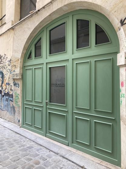 An over-sized, curved green entrance to a studio on a small monochrome side street of Paris reframes a normal-size green door with a glass panel featuring the words "open the door and begin your journey" in English.