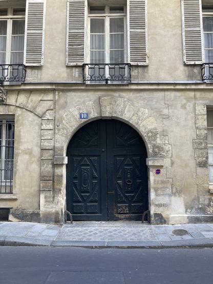 A large dark-teal blue set of curved doors addressed as number 10 set comfortably closed in a stone building with shuttered windows above and on either side, a tiny 'no parking' emblem to the right the only sign of what era this might be.