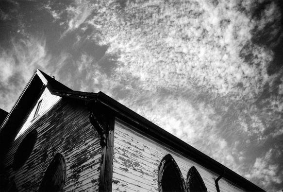 A black and white photograph of an old church building with peeling paint. Shot upwards, the steeple and top of the front and side are visible, with a sky dotted with clouds.

Ilford SFX, a near-infrared film was used with a red filter, so the shot is moody, with a lot of contrast between the shadowed and sunlit areas.