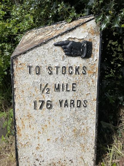Cast iron mile marker painted white. Some rusting showing through. Black lettering reads ‘TO STOCKS 1/2 MILE 176 YARDS’. Gorgeous black manicle at top pointing left. Hedgerow behind.