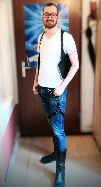 A young man wearing jeans, a leather leg harness, boots and a leather bar vest over a white t-shirt.