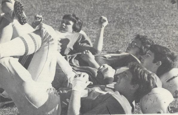 Black and white photo of four football players, lying on the ground, heads resting on helmets, legs crossed.