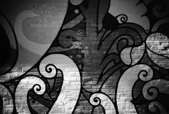 A black and white photograph, taken with near-infrared film, showing a portion of a mural painted on a brick wall. The pattern shows stylized botanical shapes, and to the left there’s a swan’s neck, the head looking back to the right. Around the swan are small stars and musical notations with the phrase “Music is  Life…”