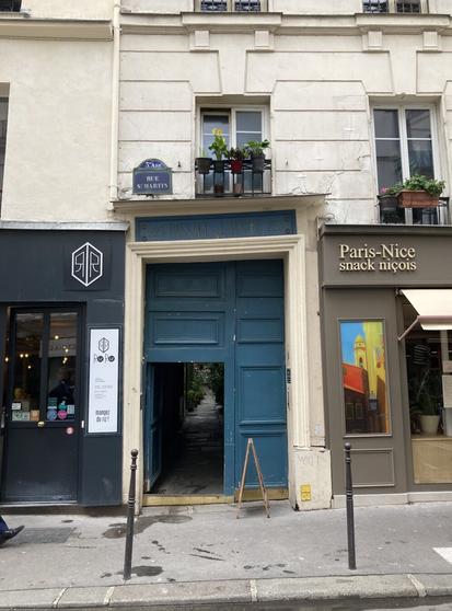 A very crooked off-white, beveled door frame on a small Paris street between two restaurants surrounds a one and a half sized door jam as if it's been pasted on in an after-thought, a too small to be a regulation sized blue door panel on the left revealing a mysterious interior garden path behind it.