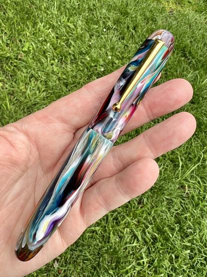 Colorful Zephyr pen model, capped, in maker’s hand, against a green grass background.