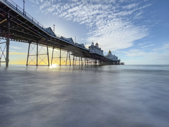 Eastbourne pier from below early in the morning.  The sea is smooth and a yellow sun is reflected off the sea under the pier.