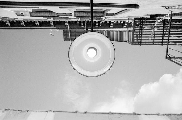 A black and white photograph, taken in the daytime, looking straight up at the wall of a brick building. The primary focus is a circular overhead light protruding straight out from the building, a lit bulb in the center. Building windowsills and fire escapes are behind it, higher on the wall. The sky is dotted with clouds.

The roof line of the adjacent building on the other side of the alley crosses the bottom of the frame.