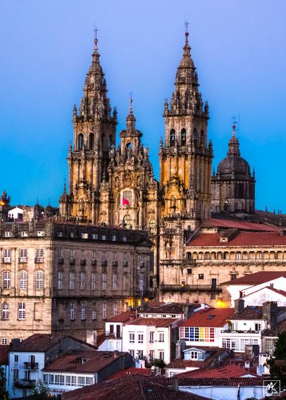 Color photo of a cathedral lit by the setting sun and rising above white buildings with red tile roofs in the town of Santiago de Compostela. The cathedral facade has two tall towers with spires on either side of a lower central spire and behind those can be seen a domed tower.