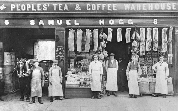 Black and white photo. Six men in aprons, one holding horse by bridle stand in row outside The Peoples’ Tea and Coffee Warehouse (Samuel Hogg).