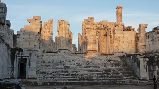 Within the sacred space of a Greek temple, a broad monumental staircase leads to the entrance to the temple. A doorway open to the priestly realm. in the pronaos, Ionian columns rise to a blue sky