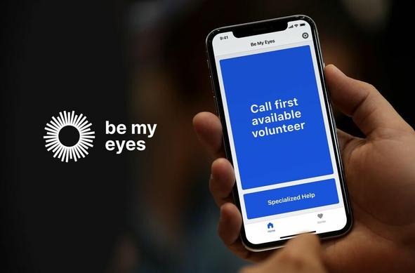 The picture is of a hand holding a smartphone with the Be My Eyes app open on the screen. There are two options on the screen, one says "Call first available volunteer" and the other says "Specialized Help".