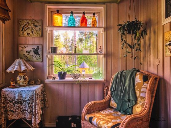 A small home entry room with an arm chair and small lace covered table and lamp. A three-shelf window is centered in the photograph. The top shelf has four colored bottles, the middle shelf has a candlestick along with a few knickknacks and sill has a small potted plant, a bowl, a pitcher of water, and a handfull of sea shells.