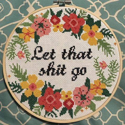 A cross stitched wreath of red, pink, and yellow flowers with light and dark green leaves. In the center are the words “Let that shit go” in cursive.It is displayed in a wooden hoop and is laying on a turquoise and cream pillow.