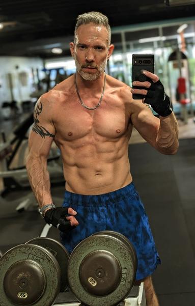 Gym mirror selfie, shirtless, standing in front on a metal dumbbell rack, wearing blue BIKE gym shorts and black lifting gloves, sweaty, slightly flexing my left arm making those arm veins bulge