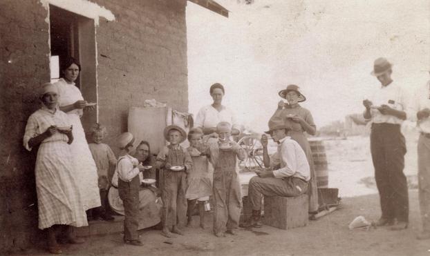 Real photo post card of farm family having dinner in front of their sod house.