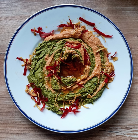 Auf einem Teller zwei Sorten Hummus, eine hell, die andere grün, sie sind wie ein Strudel ineinander gerührt, man sieht beide Farben noch. In der Mitte eine Vertiefung mit Öl. Auf dem Hummus frittierte Streifen Ingwer und rote Chili + Knoblauchscheiben.