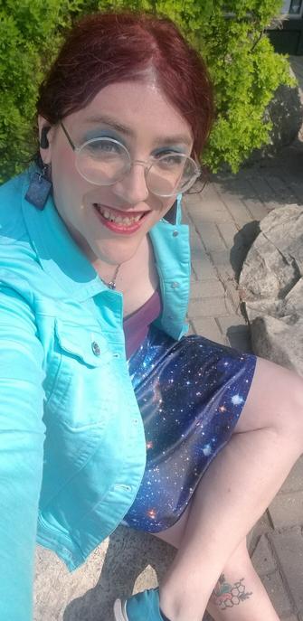 Selfie of a redheaded woman with round glasses wearing a light green jean jacket and galaxy print skirt, sitting on a rock