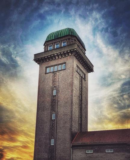 Colourful picture of an old water tower (built in 1924) with green-shimmering copper roof, with fiery sunset colours in the background.
