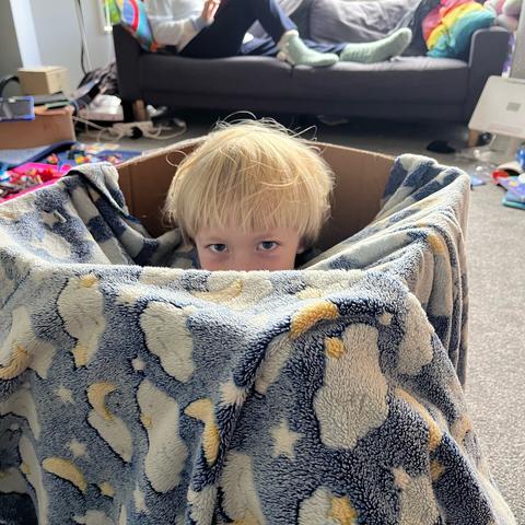 A small blonde child crouched inside a box with a fluffy blanket.