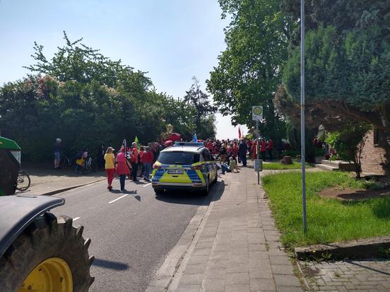 Menschen in roter Kleidung auf Straße in Dorf. Links ein Treckerreifen. Davor steht ein Polizeiwagen.